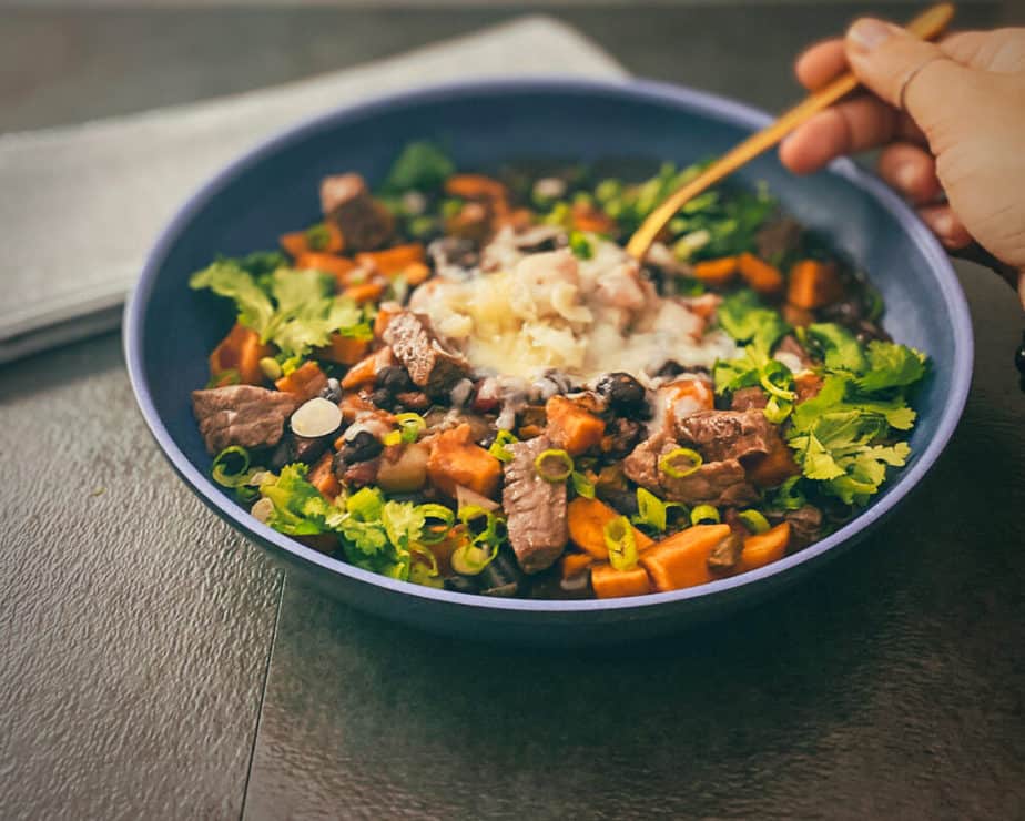 Steak chili with a hand holding a gold colored spoon and a grey and white napkin on a grey table with a blue bowl.