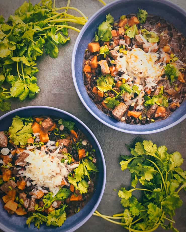 Another overhead shot of steak chili in blue bowls with white cheddar cheese, green onions, cilantro and hot sauce, and two bunches of cilantro on either side