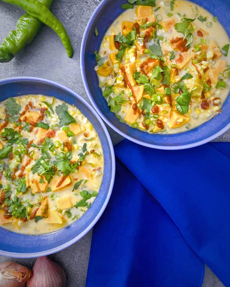 Overhead shot of two bowls of my Corn and Sweet Potato Tom Kha Soup in blue bowls with a bright blue napkin, mesilla peppers, and red onions