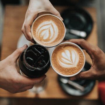 overhead shot of 3 hands cheersing with lattes and iced coffee. photo credit nathan dumlao on unsplash.