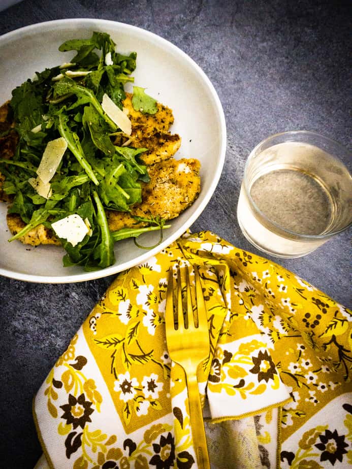 Overhead shot of chicken paillard dinner with arugula salad, glass of pinot grigio and yellow floral napkin on concrete table
