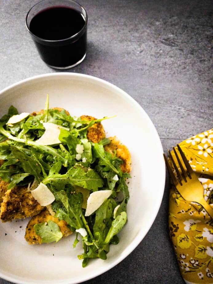 Overhead shot of chicken paillards topped with arugula salad, with glass of pinot noir, gold fork and retro 70s yellow floral napkin on grey table