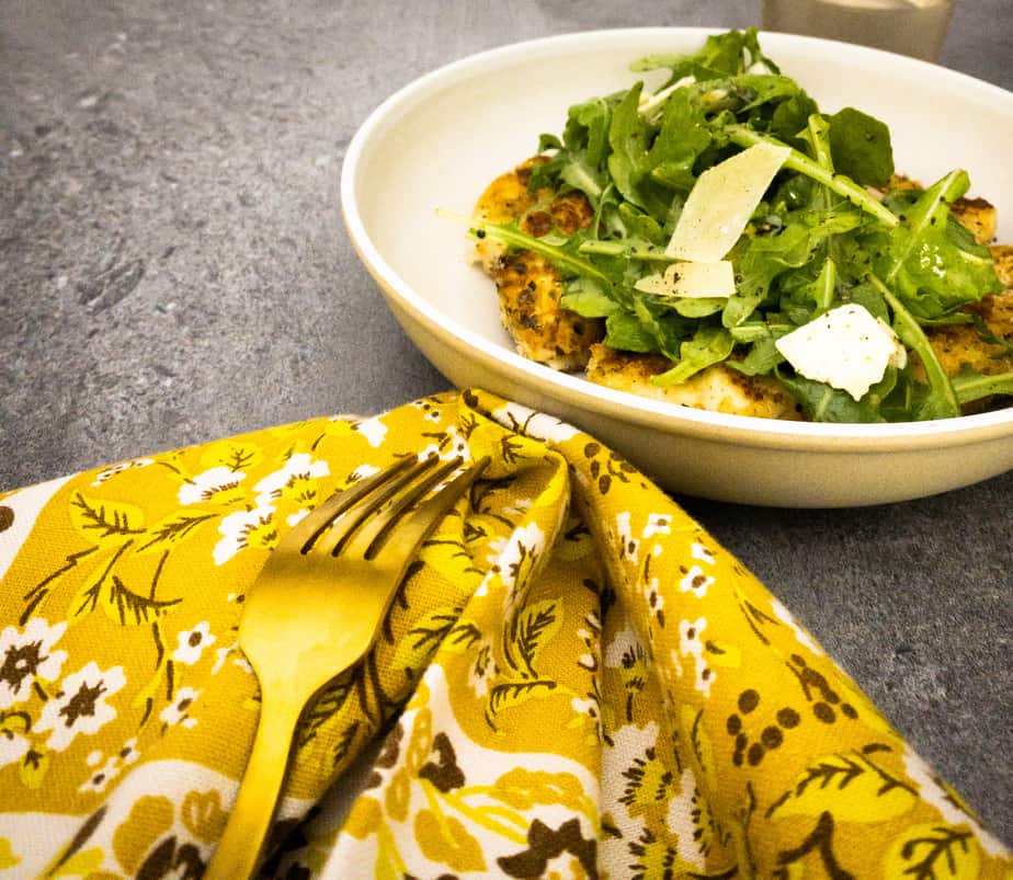 Final shot of retro 70s yellow floral napkin and gold fork pointing towards a pale neutral pasta bowl with chicken paillards topped with a simple lemon arugula parmesan salad