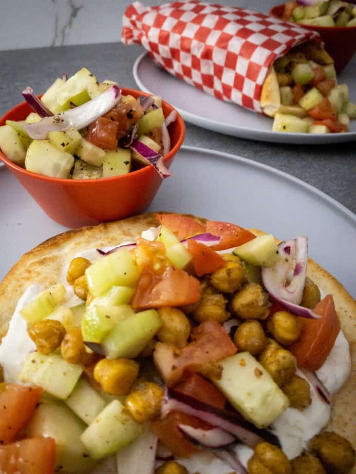Close up shot of Chickpea Gyros with Garlic Yogurt and Chopped Tomato, Cucumber and Red Onion Salad. In the background, one of the gyros is wrapped in a red and white check paper with extra salad in a red bowl on the plate.