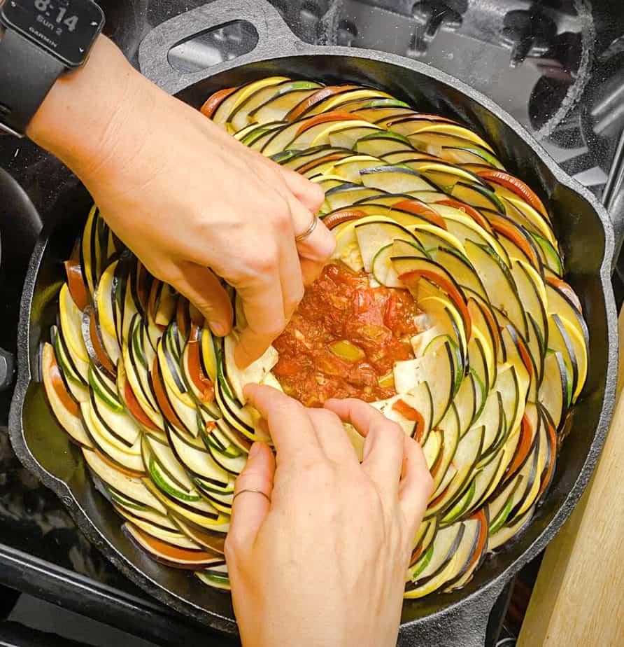 Overhead shot of me assembling the vegetables for the ratatouille in a cast iron skillet. You can still see the tomato-bell pepper sauce in the middle of the pan where I haven't finished arranging vegetables.