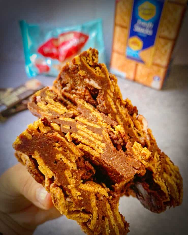 A hand holding a stack of three indoor s'mores bars with traditional s'mores ingredients in the background