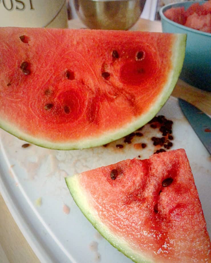 Close up shot of a quarter of a bright red watermelon on a white cutting board with a smaller piece of melon closer to the frame, seeds are scattered on the board where they have been removed.
