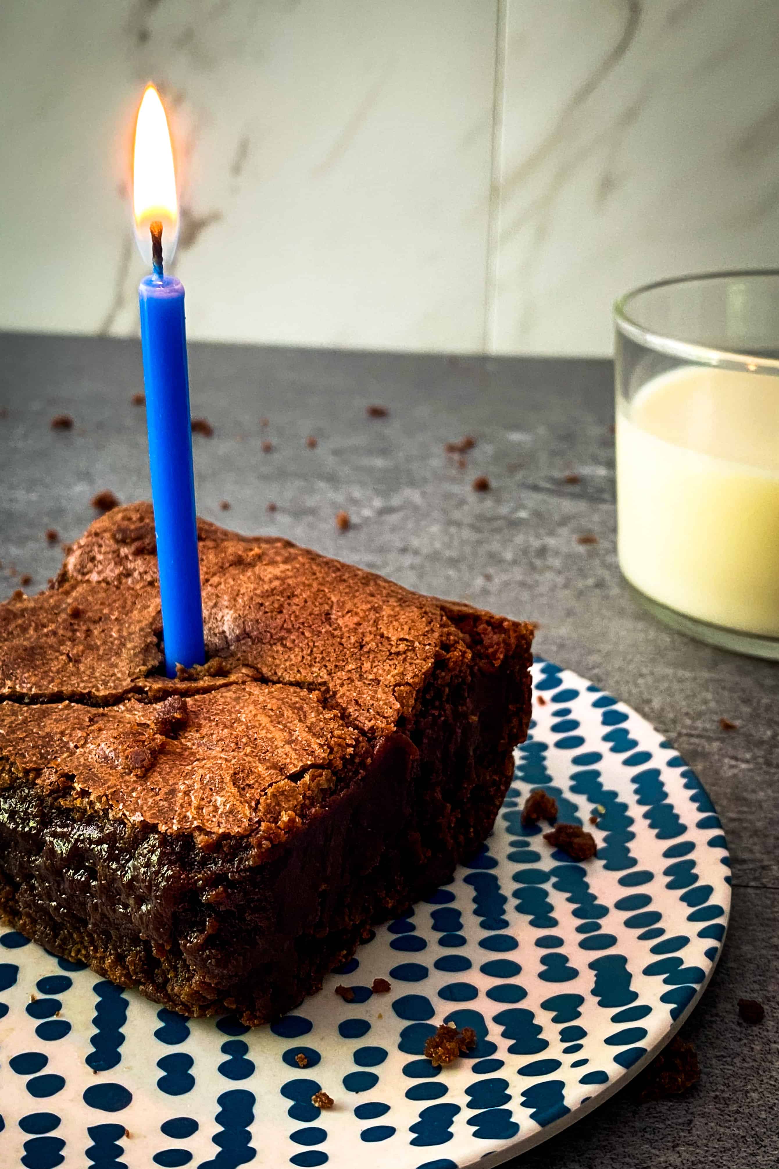 One of Julia's favorite fudgy brownies on a small blue and white plate with a blu candle lit for her birthday, and a glass of almond milk in the background.