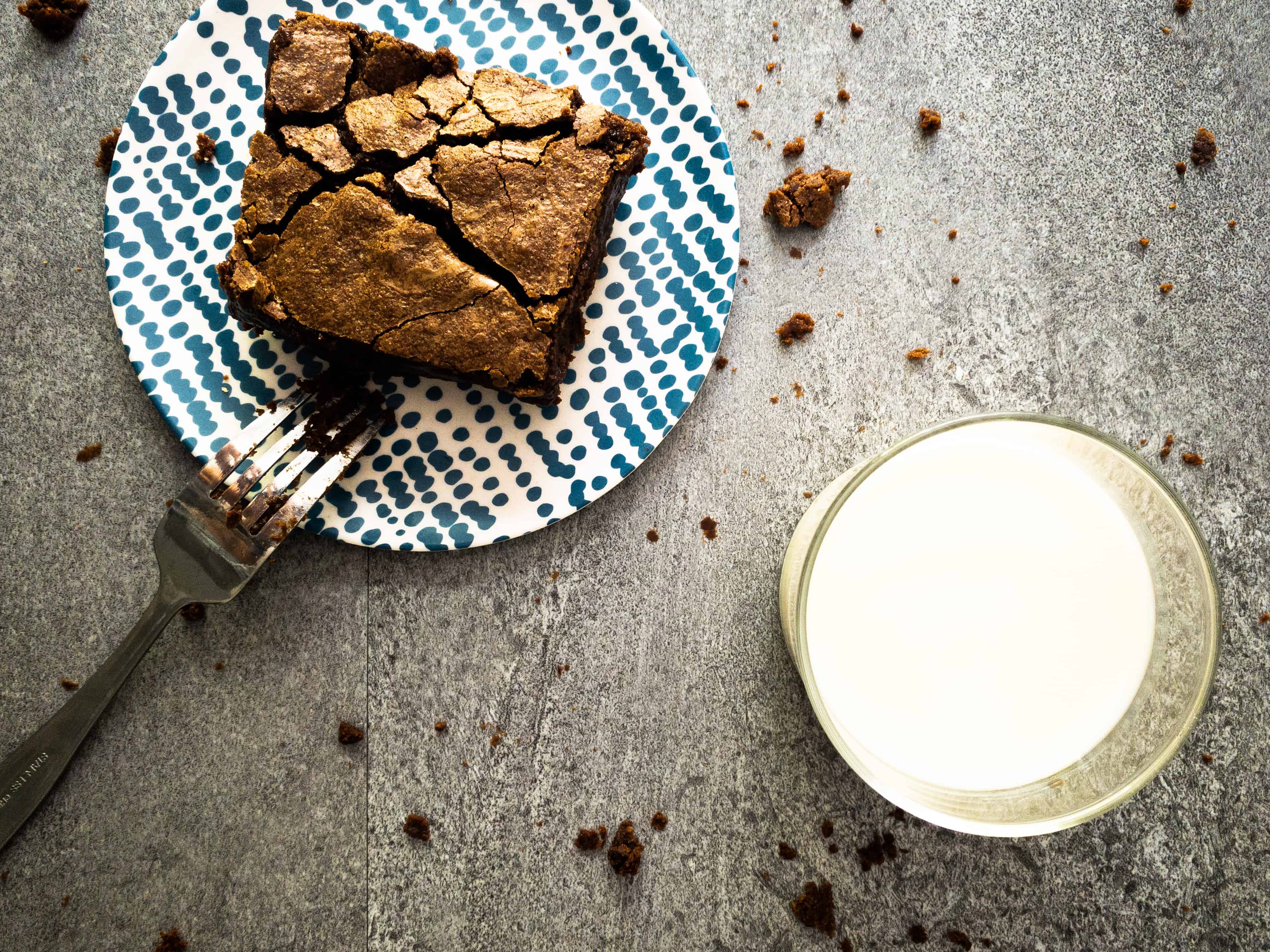 Overhead shot of brownie on a plate with a glass of milk. The fork is covered in fudgy particles sticking to it