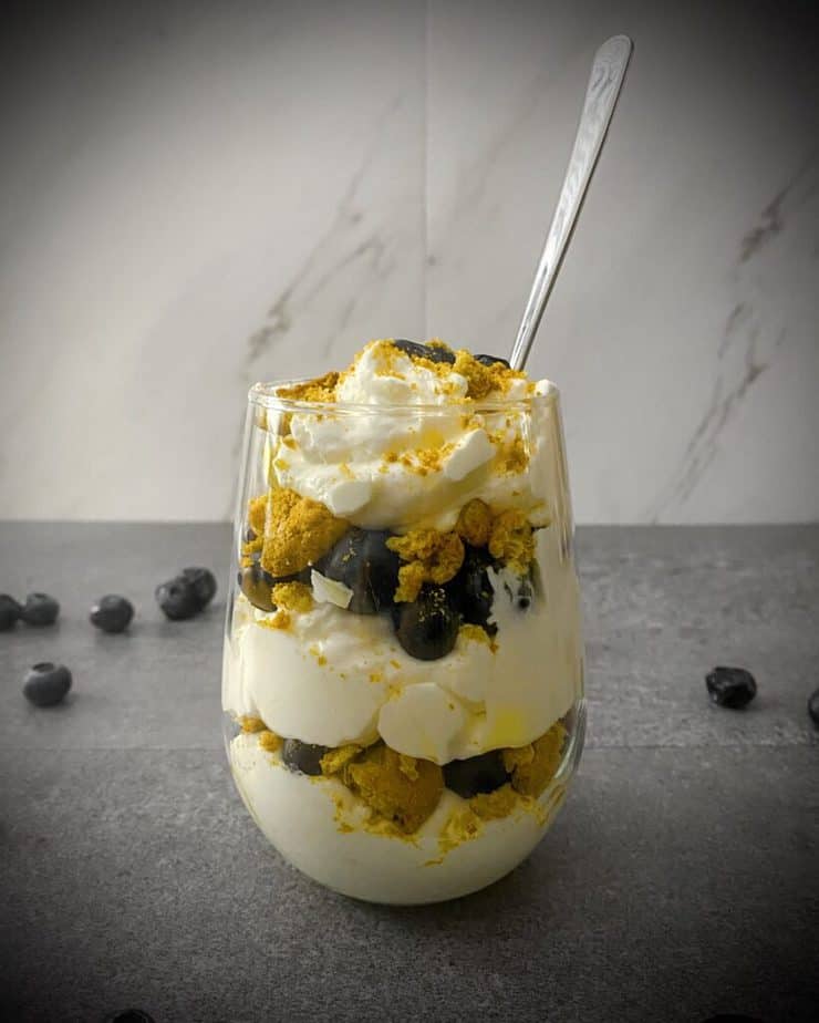 Close up sideways shot of lemon blueberry ginger trifle in a stemless wineglass with a silver spoon sticking out of it and blueberries scattered around