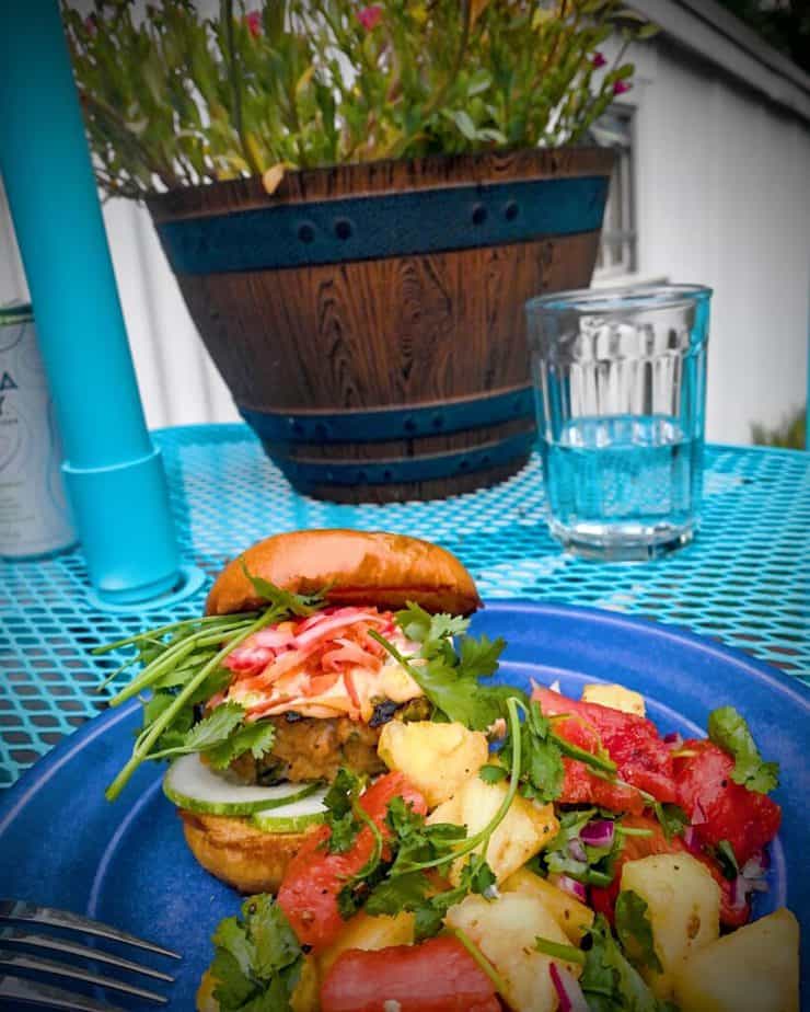 Outdoor shot of a blue plate with a pork burger and spicy pineapple watermelon salad on a turquoise wire table with a pot of pink and yellow flowers in the back.