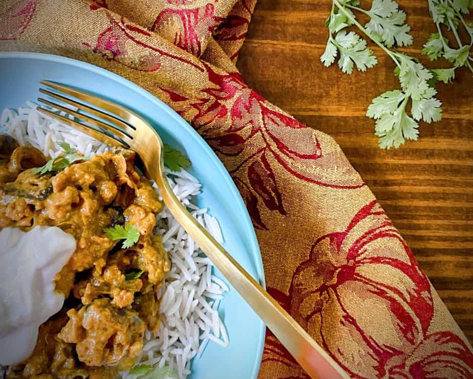 cauliflower tikka masala over basmati rice garnished with yogurt in a turquoise bowl with a gold fork on a wooden table with a maroon and orange dish towel
