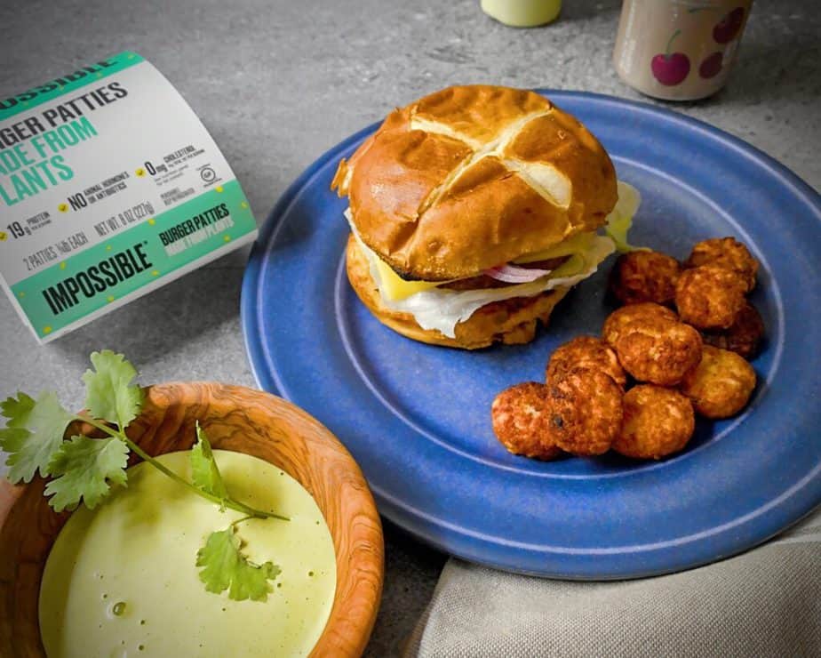 Peruvian Green Sauce in a wooden bowl next to a blue plate with an impossible burger on a pretzel bun and tater tots