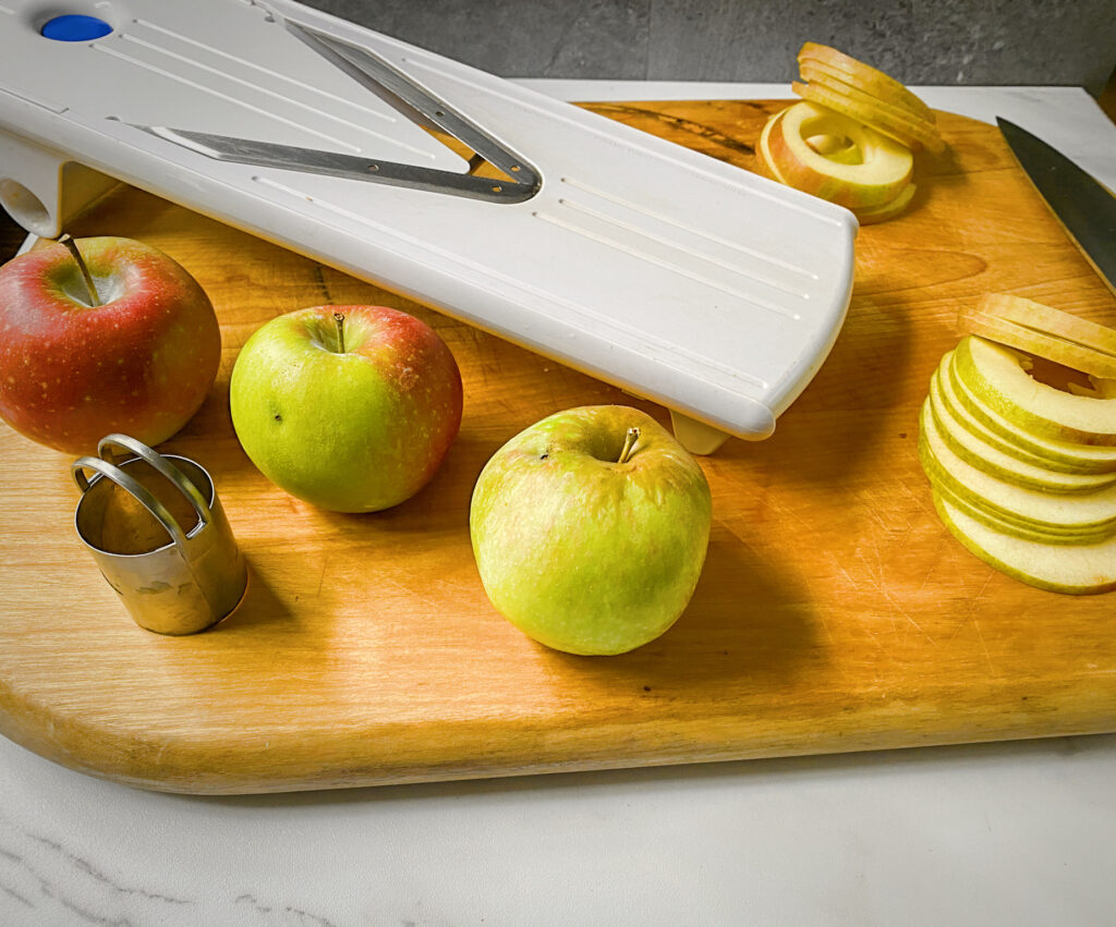 mandoline on a wooden cutting board with three unsliced apples and two sliced apples