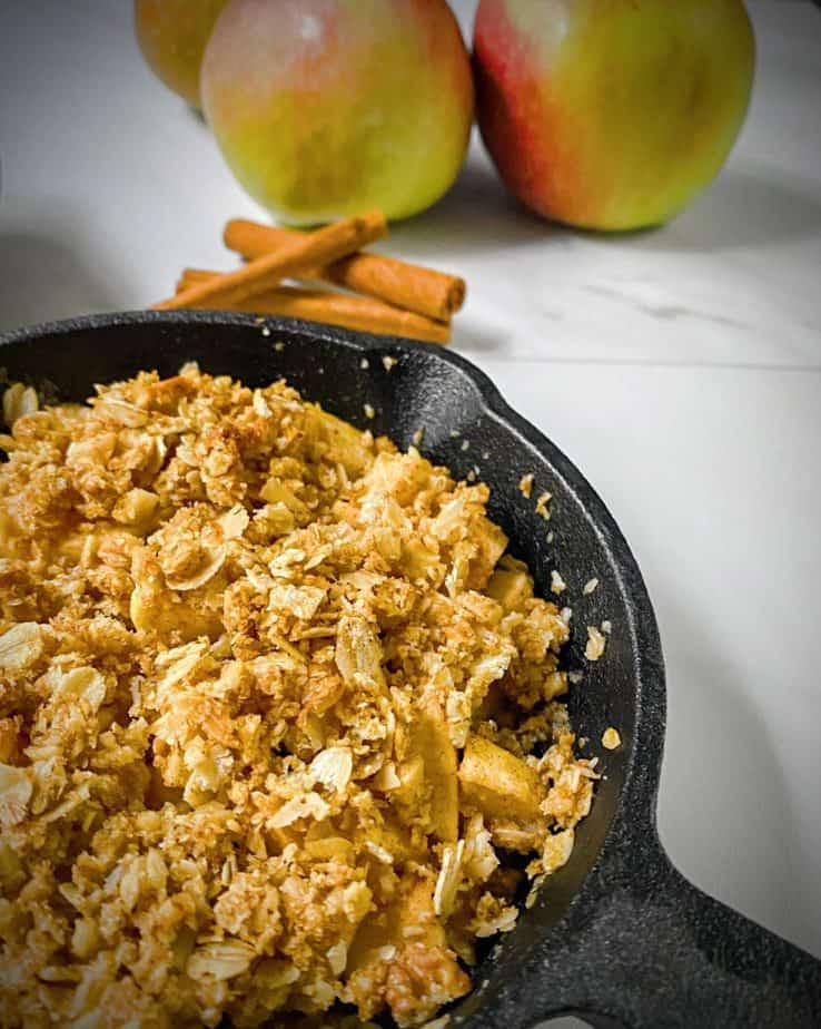 mini apple crisp for two in a cast iron pan with 3 apples and cinnamon sticks in the background