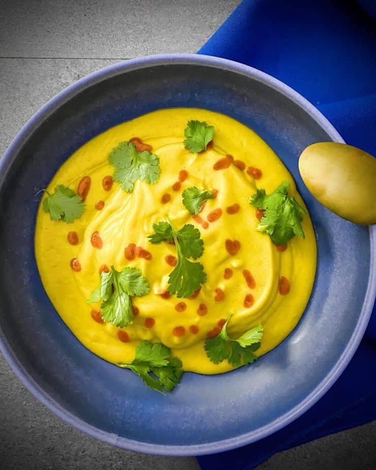 overhead shot of spicy roasted butternut squash and apple soup in a blue pasta bowl garnished with sriracha and cilantro, with a gold spoon and a bright blue napkin