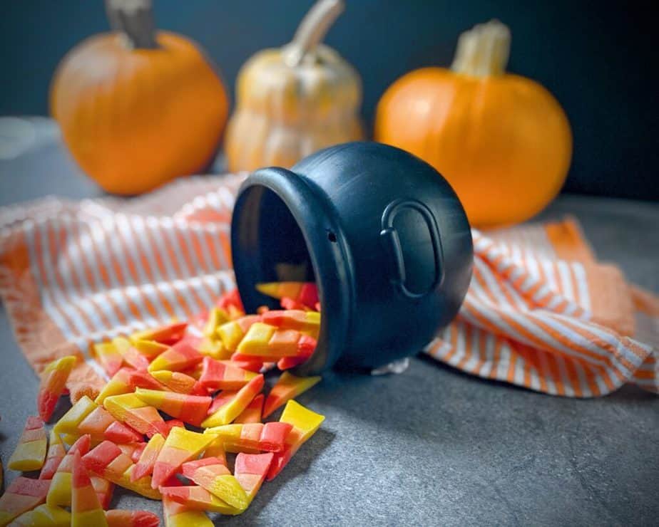 homemade yellow, orange and red candy corn spilling out of a plastic cauldron on a orange and white striped tea towel with three pumpkins behind on a black background