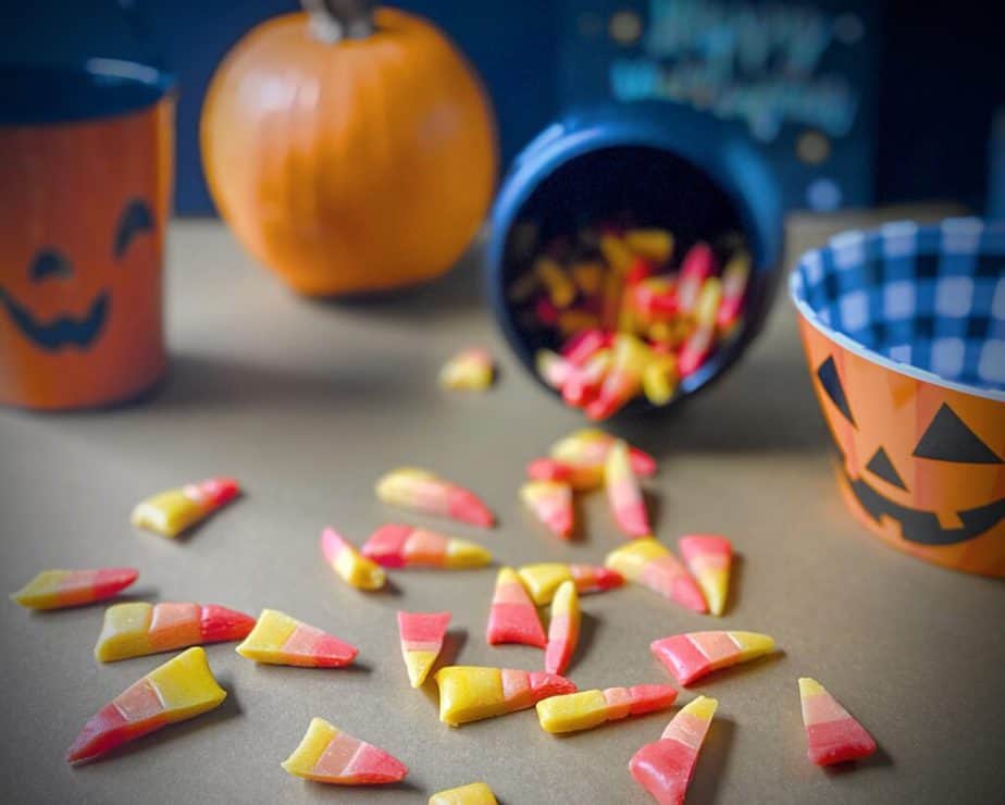 red, orange and yellow striped candy corn made with honey spilling out of a black plastic cauldron surrounded by jackolanterns and a full pumpkin on a gold table with a black background