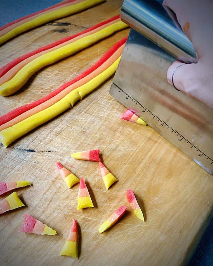 hand holding a bench scraper cutting rainbow ropes of homemade candy corn on wooden cutting board