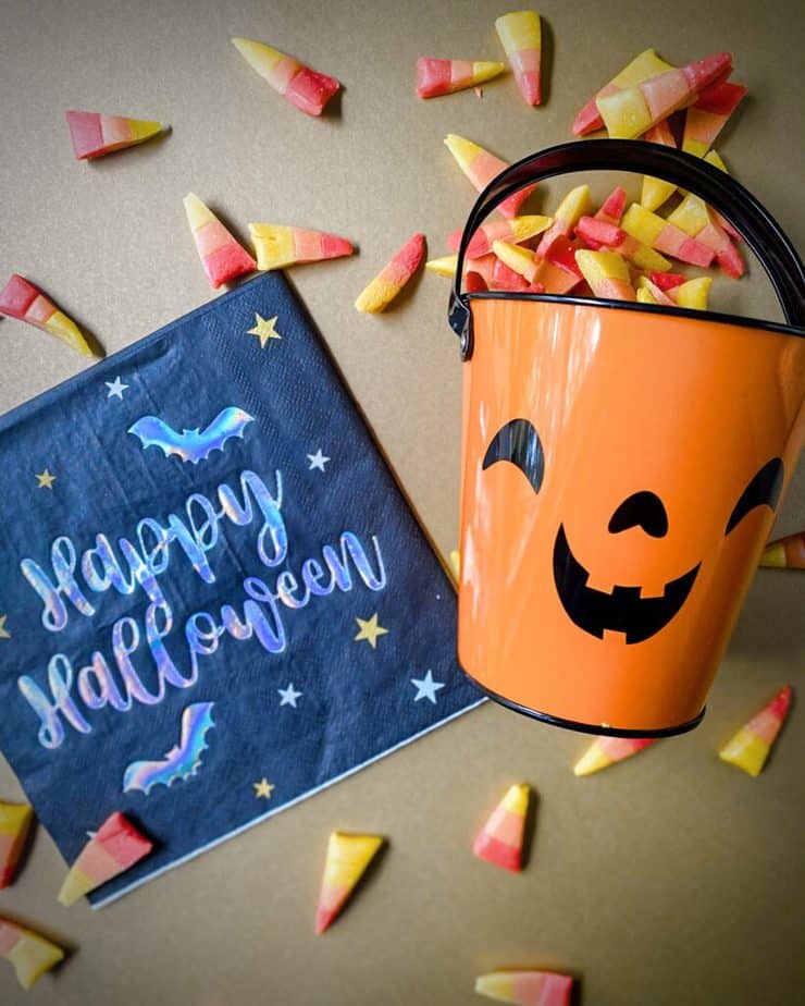 red, orange and yellow candy corn spilling out of a jack o lantern designed candy bucket on a gold background with a black, irridescent and gold napkin saying "happy halloween"