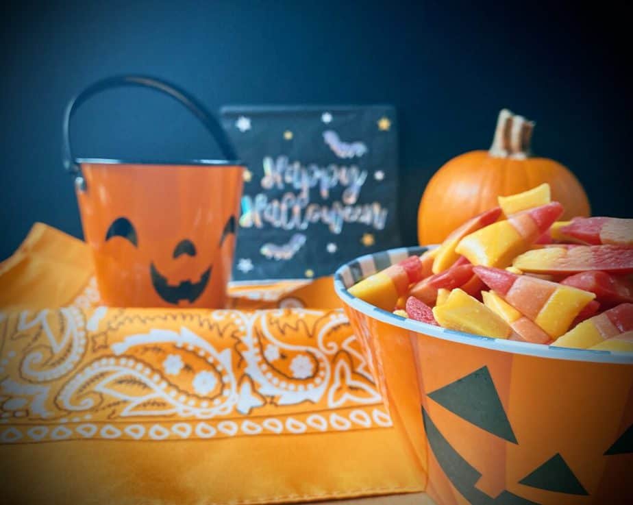Close up of a Jackolantern treat bowl filled with homemade candy corn on an orange handkerchief print with a black background with a jackolantern shaped bucket, sugar pumpkin and irridescent happy halloween sign behind it