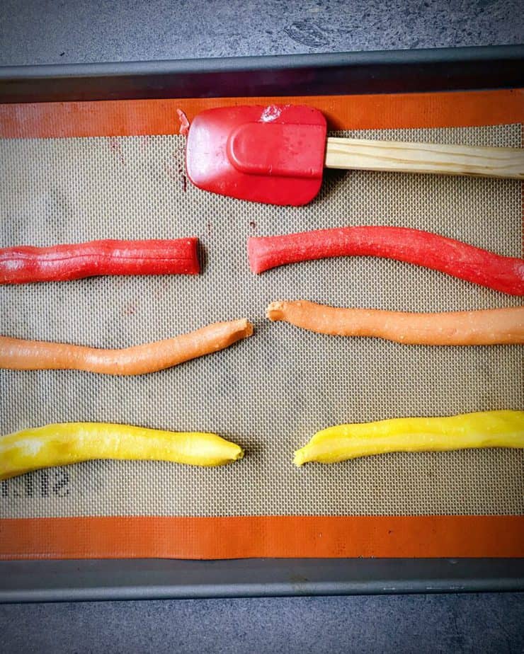 six strands of homemade candy corn dough made with honey - 2 each red, orange, and yellow on a silpat sheet with a red silicone spatula