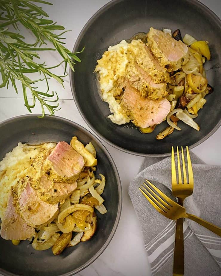 overhead shot of two pasta bowls with finished mustard glazed pork with apples, fall veggies, mashed potatoes and pan gravy with fresh sprigs of rosemary, gold forks and a light grey linen on a white marble background