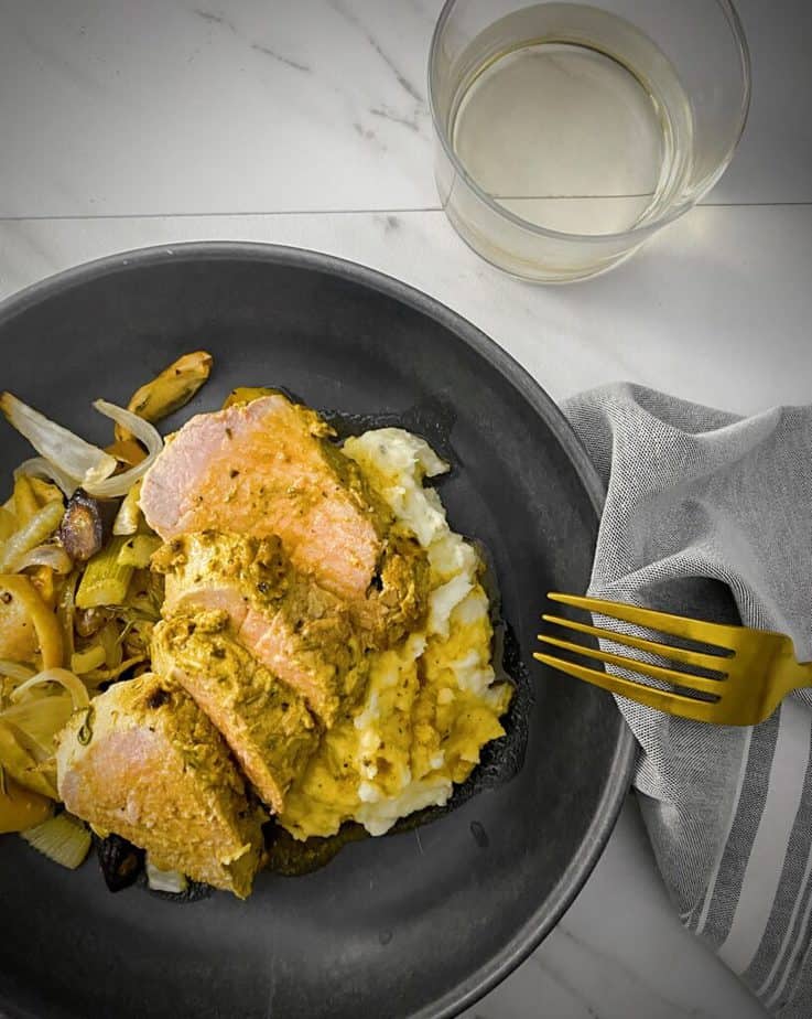 overhead shot of mustard crusted pork tenderloin slices plated on a bed of roasted apples and root vegetables, mashed potatoes and topped with pan gravy in a dark grey pasta bowl with a gold fork, a glass of white wine, and a light grey napkin