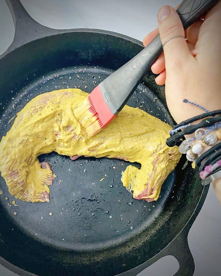 hand holding a pastry brush brushing on mustard mixture on pork tenderloin in a cast iron skillet