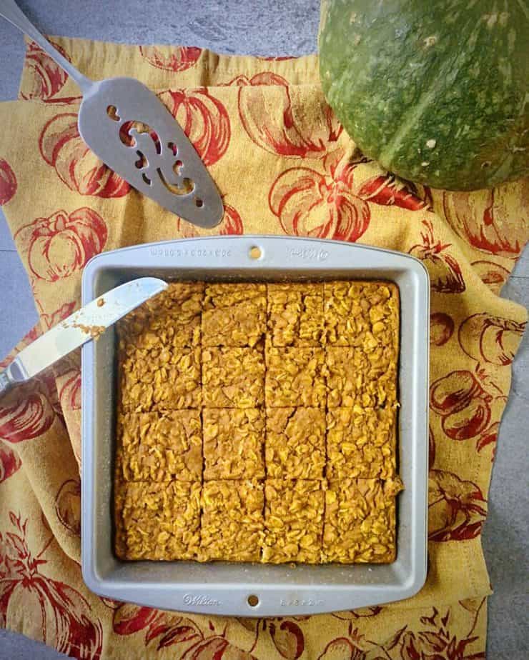 overhead shot of pumpkin baked oatmeal in a square tin cut into 16 squares on a gold and maroon pumpkin themed tea towel 