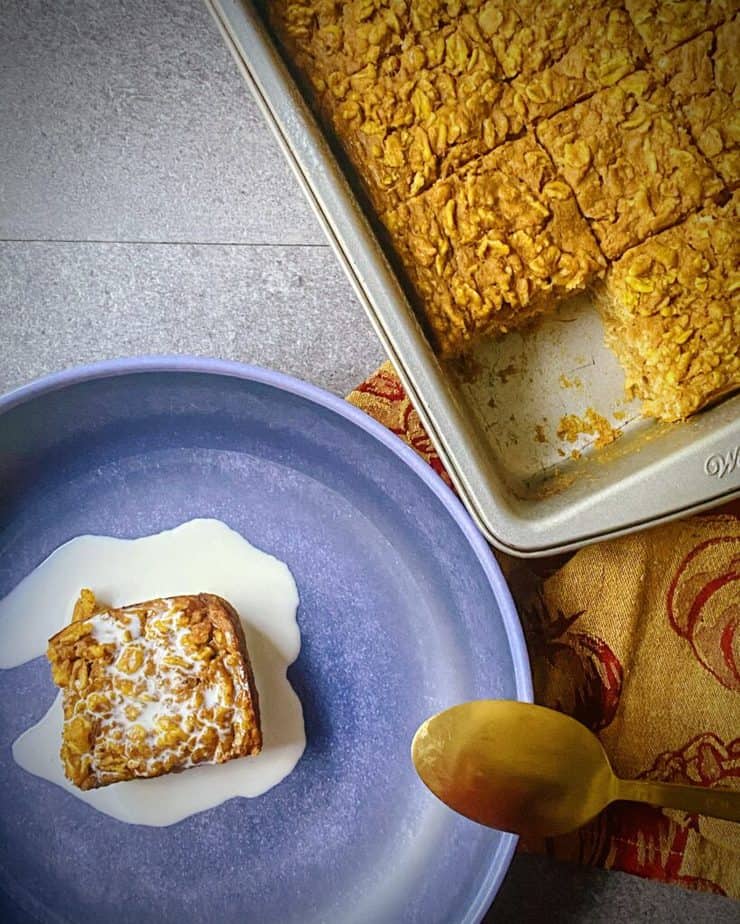 overhead shot of one slice of pumpkin pie baked oats removed from the square tin next to a blue pasta bowl with the slice covered in cream and a gold and maroon pumpkin themed tea towel