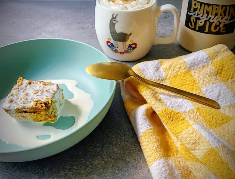 breakfast scene with baked oatmeal topped with cream in a turquoise bowl with a gold spoon, a yellow and white gingham tea towel, a llama patterned mug with a coffee drink topped with whipped cream and cinnamon sugar, and a jug of homemade pumpkin spice syrup