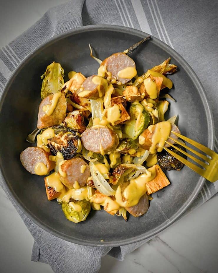 finished dish of sheet pan sausage and fall veggies drizzled with creamy dijon apple vinaigrette in a grey pasta bowl on a grey napkin with a gold fork