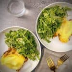 two plates of lamb and eggplant moussaka shepherd's pie with arugula salad in white pasta bowls on a grey slate background with a glass of sparkling water and gold forks