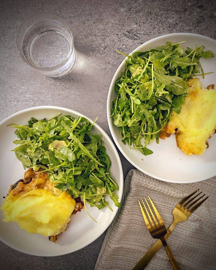 two white pasta bowls with lamb and eggplant shepherd's pie and arugula salad on a grey slate background, gold forks and a tan waffle knit napkin