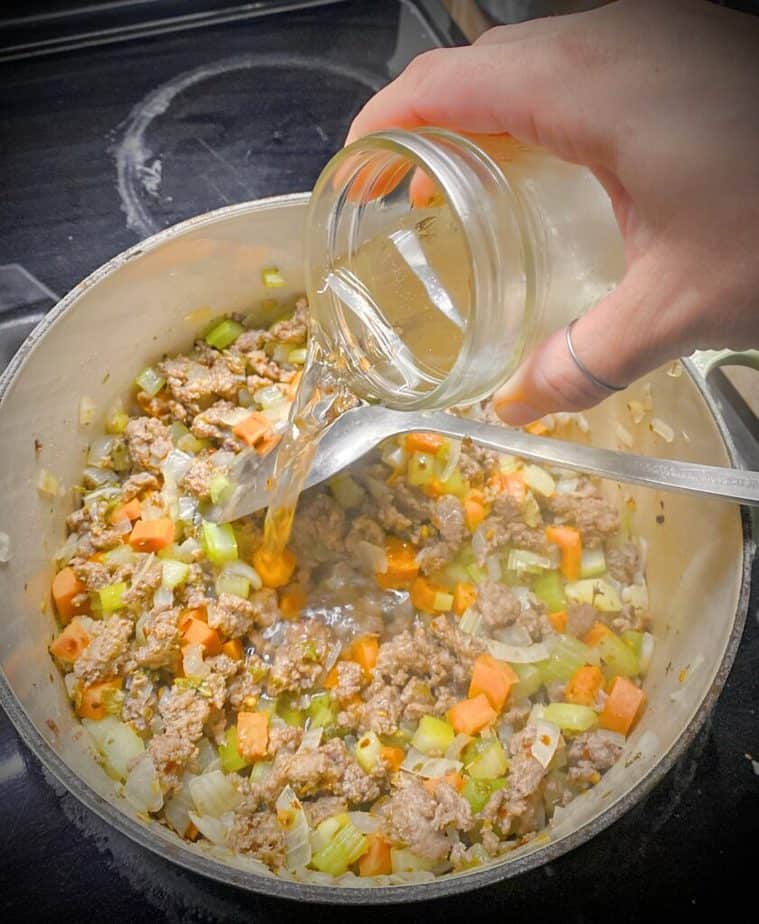 hand pouring white wine into dutch oven with lamb and mirepoix for lamb and tomato bolognese sauce