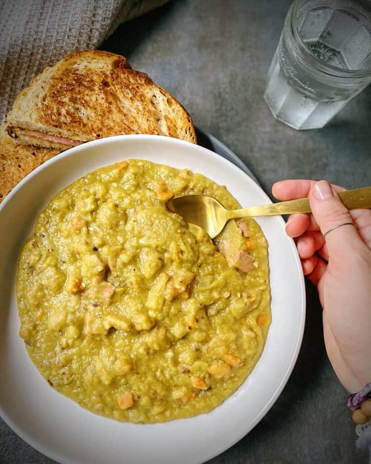 hand holding a gold spoon dipping into a bowl of split pea and ham soup with a smoked gouda and ham grilled cheese sandwich and a glass of sparkling water on a grey slate table