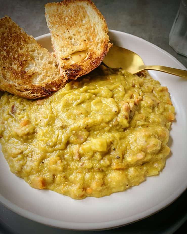 close up shot of split pea and ham soup in a white pasta bowl with a gold spoon and two halves of a ham and gouda smoked cheese on sourdough resting on the edge of the plate