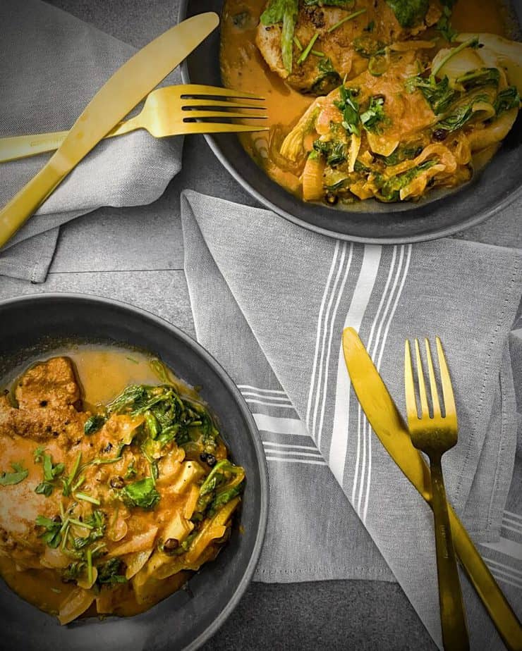 Overhead shot of two grey pasta bowls filled with chicken tagine from sun basket. grey and white striped napkins with gold flatware round out the scene
