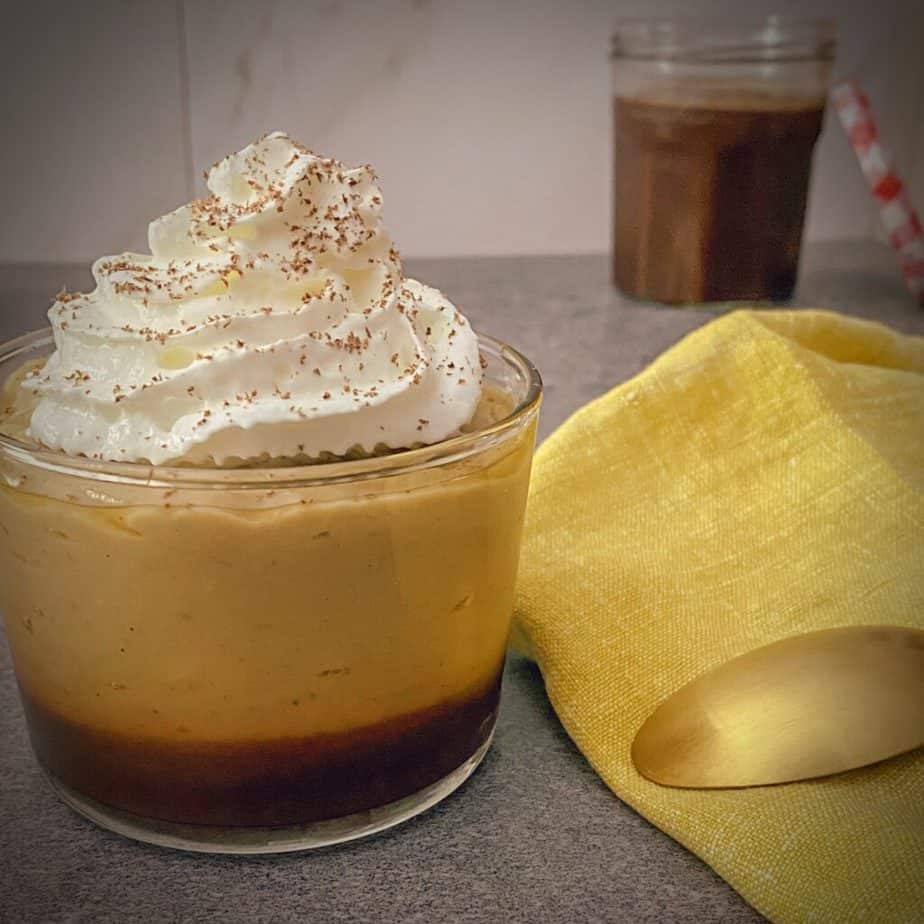 black bottomed pudding cup with layer of coffee rum fudge, homemade banana pudding and whipped cream topped with chocolate shavings in a clear cup on a grey surface with a yellow napkin and gold spoon