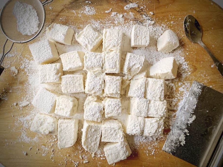 homemade marshmallows turned out on a wooden cutting board after being cut into cubes with a bench scraper and corn starch