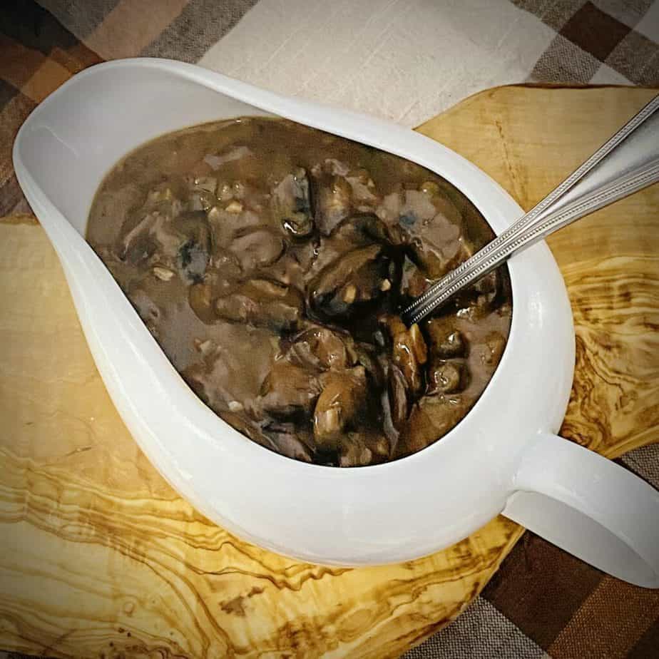 overhead shot of mushroom gravy in a white gravy boat on a piece of olive wood with a silver ladle spoon and a light orange, brown and white plaid napkin