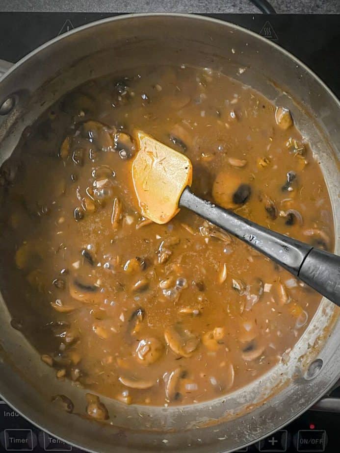final shot of mushroom gravy in saucepan with red and black silicone spatula. sauce is visibly thickened and darkened after cooking down.