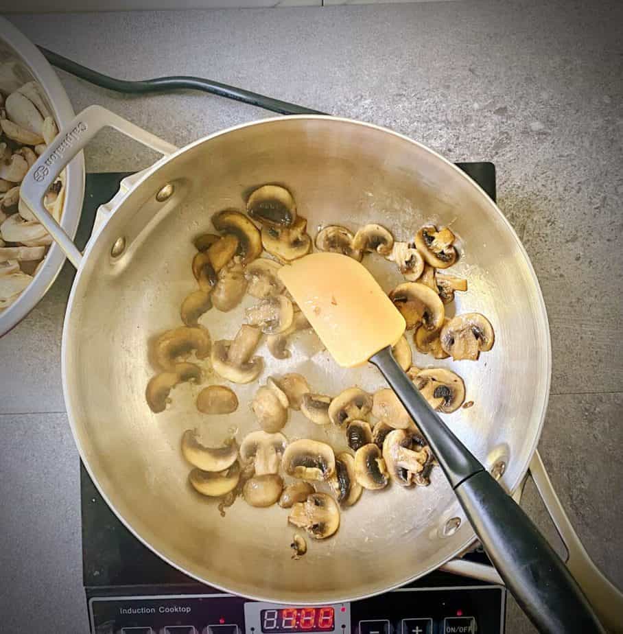 first batch of mushrooms that have been properly sautéed to a golden brown in saucepan with remaining raw mushrooms in a bowl to the side