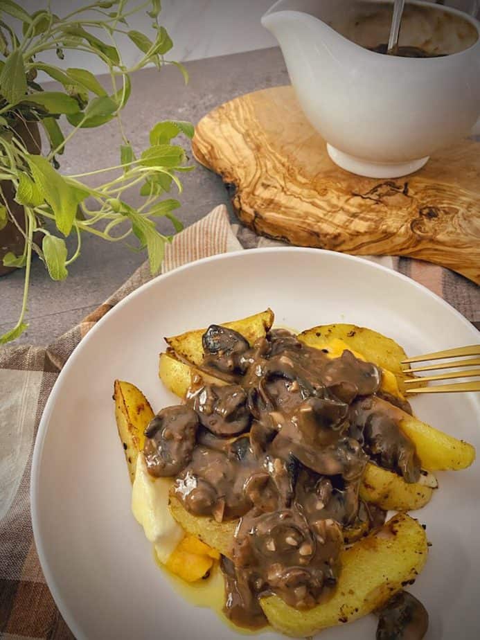 final shot of completed poutine recipe in a white pasta bowl on a light orange, brown and white tea towel with a gravy boat on a piece of olive wood and a sage plant in the background