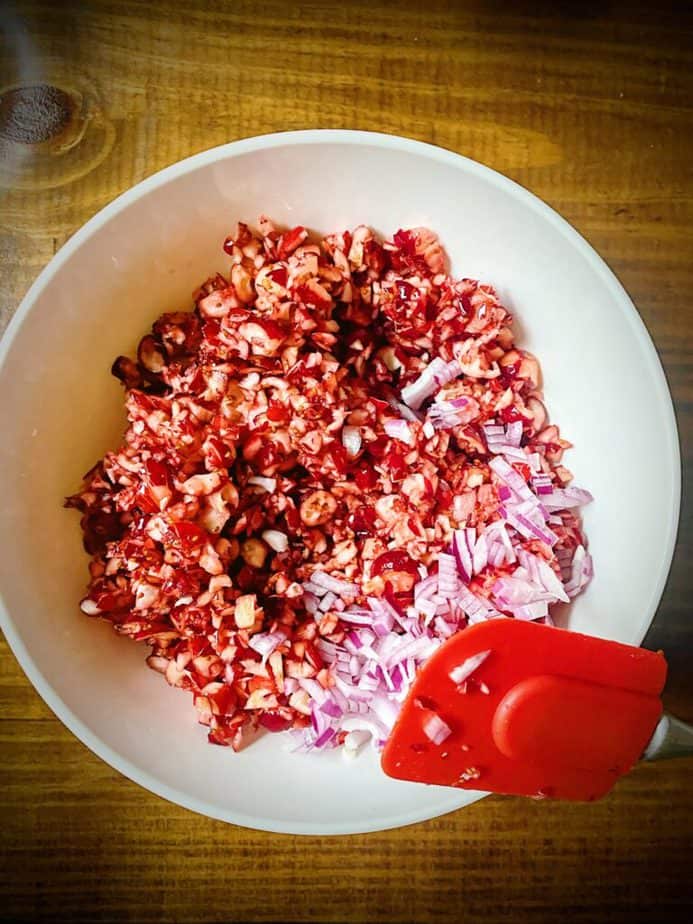 processed cranberries in white bowl with minced red onion on a wooden table with a red spatula