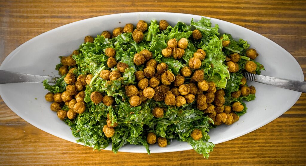 vegan kale salad with tandoori spiced crispy chickpeas and a roasted garlic tahini dressing in an oblong white bowl with silver salad spoons on a wooden table