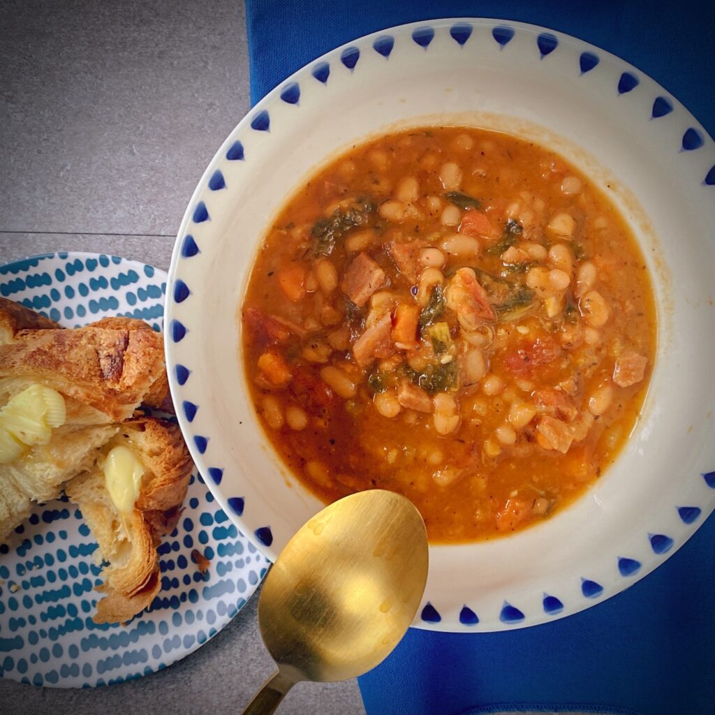 Instant Pot Navy Bean Soup with tomatoes, kale and ham in a white soup bowl with a blue border and some buttered croissant toast on a navy and white dotted plate with a gold spoon.