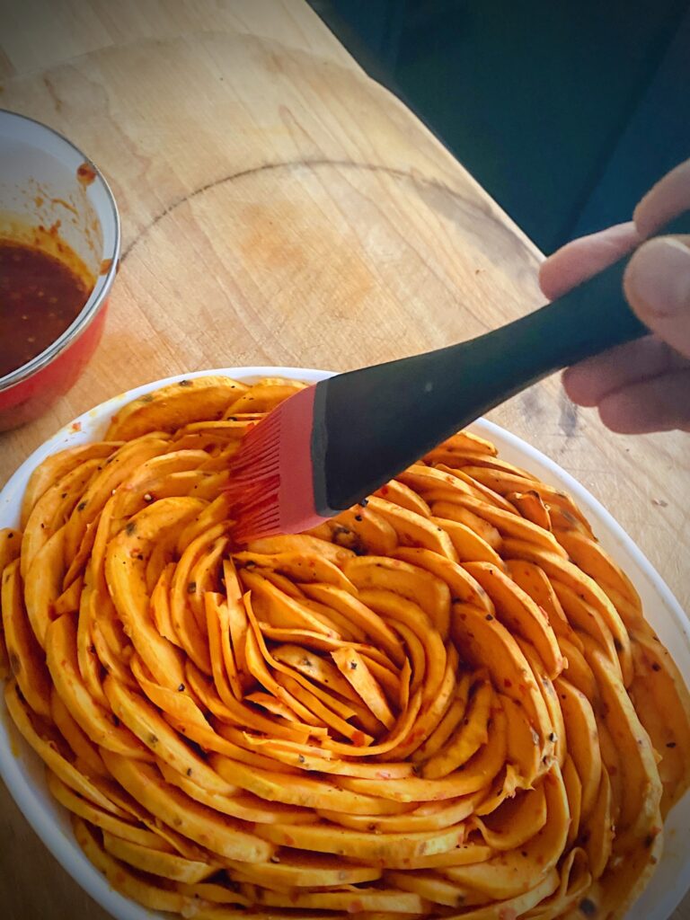 hand holding pastry brush that is brushing the top of the sweet potato tian with maple harissa sauce mix