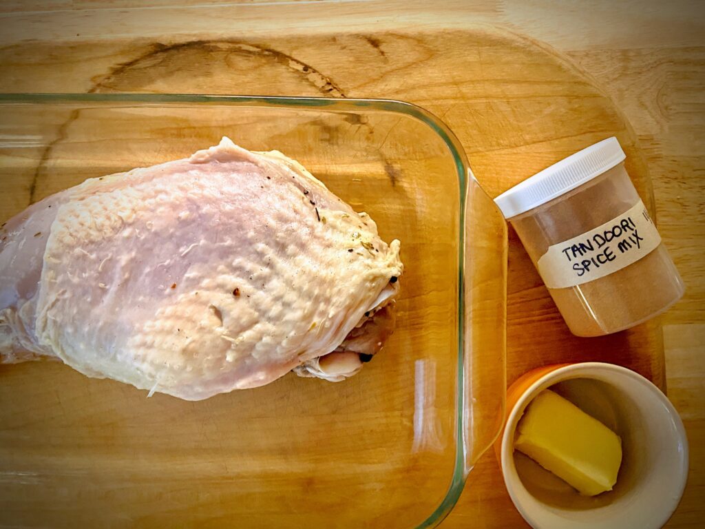 brined turkey breast in a roasting pan on a wooden cutting board with a jar of homemade tandoori spice mix and a small bowl with softened butter to the side